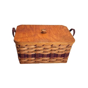 May include: A rectangular woven basket with a wooden lid and leather handles. The basket is made of natural brown wicker with purple accents. The lid has a wooden knob.