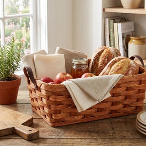 May include: A woven brown basket filled with fresh bread, apples, and a jar of honey. The basket also contains folded linen napkins and has leather handles. A small potted rosemary plant sits nearby.
