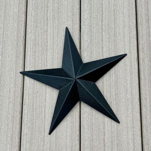 May include: A black, five-pointed star with a textured surface. The star is centered on a light gray wooden background. The star's points are sharp and well-defined, creating a bold, rustic aesthetic.