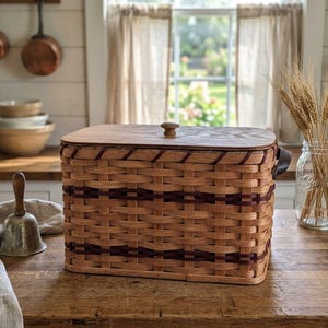 May include: A rectangular woven basket with a flat wooden lid and a small wooden knob handle. The basket features a natural light brown finish with two horizontal bands of dark purple woven material and a dark leather side handle.