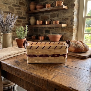 May include: A rectangular woven basket with a burgundy and natural color pattern, featuring leather handles. The basket sits on a rustic wooden table, alongside a loaf of bread, pottery, and a vase of lavender.