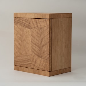 May include: A small wooden cabinet with a single door. The cabinet is made of light brown wood with a natural grain pattern. The door is closed and has a simple design.