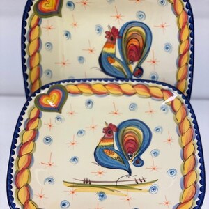 May include: Two square ceramic plates with a rooster design. The plates have a cream-colored background with a blue border. The rooster is painted in blue, red, yellow, and orange. The plates are decorated with hearts and starbursts.