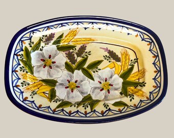 May include: Oval ceramic serving platter with a hand-painted floral design. The platter features three large white flowers with red centers, surrounded by green leaves, yellow wheat, and purple lavender. The rim is decorated with a blue and white pattern.