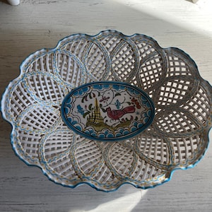 May include: White ceramic basket with a lattice design and a scalloped edge, accented with blue and yellow details. An oval dish with a bird and floral design sits inside the basket. The basket and dish are likely for decorative or serving purposes.
