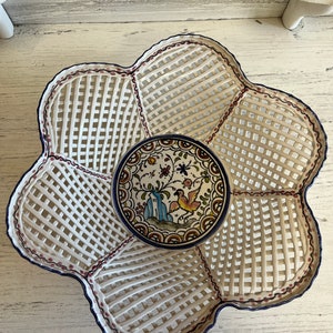 May include: A white, flower-shaped serving tray with a lattice design and a small, round, decorative plate. The plate features a colorful illustration of a bird, flowers, and a building. The tray has a dark blue trim.