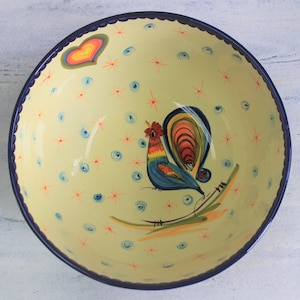 May include: A decorative ceramic bowl with a yellow interior and a dark blue scalloped rim. The bowl features a colorful rooster illustration, along with a heart and starburst pattern. The bowl is suitable for serving food.