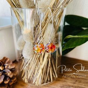 Peut inclure: Une paire de boucles d'oreilles pendantes en argent avec des perles de verre orange, rouge et blanc. Les boucles d'oreilles sont suspendues à un vase rempli de fleurs séchées. Le texte "Petite Sablaise Artisan Verrier" est visible en bas de l'image.