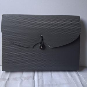 May include: A rectangular dark gray document folder with a textured, grid-like surface pattern. The folder features a curved flap closure secured by a black circular button and an elastic cord that loops around the button to keep the contents inside.