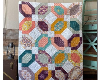 Könnte beinhalten: Ein Quiltmuster namens "Petal Junction" mit weißem Hintergrund und bunten Achteckformen. Der Quilt ist aus verschiedenen Blumenstoffen in den Farben Rosa, Lila, Türkis, Gelb und Grün gefertigt. Der Quilt wird auf einem Holzboden mit einer rustikalen Holzwand im Hintergrund gezeigt. Der Text "Highway 10 Designs" und "Fat Quarter Friendly Pattern" ist ebenfalls auf dem Bild zu sehen.
