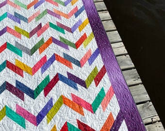 Könnte beinhalten: Eine bunte Chevron-Steppdecke mit einem violetten Rand und einem weißen Hintergrund. Die Steppdecke ist aus verschiedenen Stoffen gefertigt und hat ein gestepptes Design.