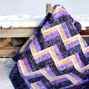 Peut inclure: Une courtepointe à motif chevron violet et or drapé sur une balustrade en bois avec de la neige en arrière-plan.