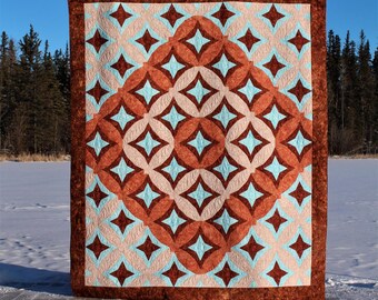 Könnte beinhalten: Eine Steppdecke mit einem geometrischen Muster aus braunen, rosa und blauen Diamanten und Kreisen. Die Steppdecke wird vor einem schneebedeckten Hintergrund gezeigt.