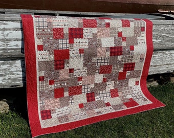 Könnte beinhalten: Eine rot-weiße Patchwork-Decke mit rotem Rand. Die Decke besteht aus verschiedenen Stoffen mit unterschiedlichen Mustern, darunter Karos, Blumen und geometrische Muster. Die Decke ist gefaltet und über eine Holzoberfläche gelegt.