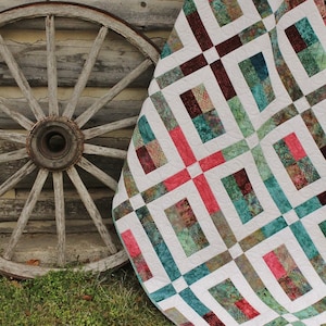 Op de afbeelding: Een wit quilt met een patchwork-ontwerp met vierkanten van teal, bruin en roze stof. Het quilt is gevouwen en leunt tegen een houten wagenwiel.