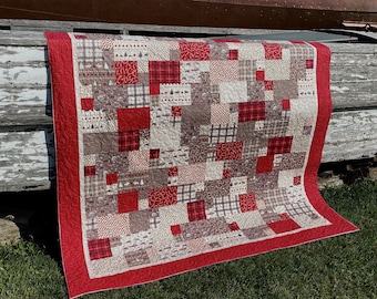 Könnte beinhalten: Eine rot-weiße Patchwork-Decke mit rotem Rand. Die Decke besteht aus verschiedenen Stoffen mit unterschiedlichen Mustern, darunter Karos, Blumen und geometrische Muster. Die Decke ist gefaltet und über eine Holzoberfläche gelegt.