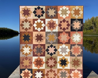 Könnte beinhalten: Eine Steppdecke mit Patchwork-Muster, die verschiedene Sternformen in Braun-, Orange- und Cremetönen zeigt. Die Steppdecke wird auf einem Holzsteg über einem Gewässer ausgestellt.