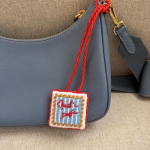 May include: A slate blue leather shoulder bag with a textured finish and gold-tone hardware. Attached to the bag is a square needlepoint charm featuring a light blue and white striped pattern with two red stylized figures, bordered by red rope trim.