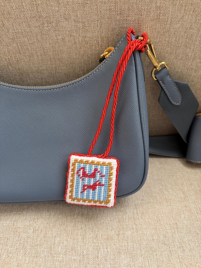 May include: A slate blue leather shoulder bag with a textured finish and gold-tone hardware. Attached to the bag is a square needlepoint charm featuring a light blue and white striped pattern with two red stylized figures, bordered by red rope trim.