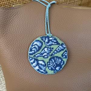 May include: A circular needlepoint ornament featuring a repeating abstract pattern in shades of dark blue, light blue, and pale green. The piece is edged with a twisted light blue cord and includes a matching light blue cord loop for hanging.