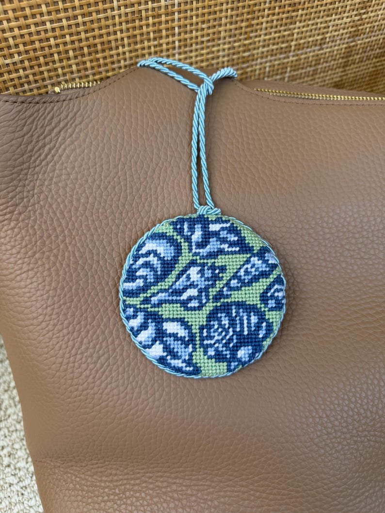 May include: A circular needlepoint ornament featuring a repeating abstract pattern in shades of dark blue, light blue, and pale green. The piece is edged with a twisted light blue cord and includes a matching light blue cord loop for hanging.