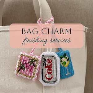 May include: Three rectangular cross-stitch bag charms hanging from a beige canvas tote. The left charm features pink and white stripes with green foliage. The center charm depicts a silver soda can with red text. The right charm shows a cocktail.