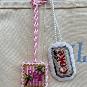 May include: Two embroidered bag charms attached to a beige canvas tote. One charm is a square featuring pink palm trees with gold accents. The other is an oval shape depicting a silver soda can with the red text Coke, both hanging by white cords.