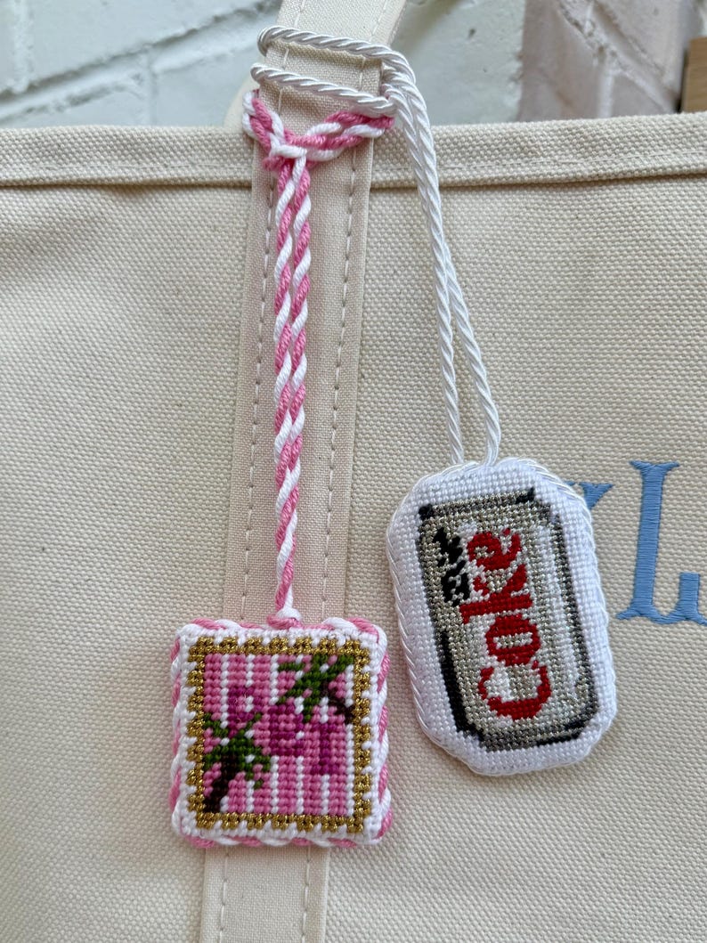 May include: Two embroidered bag charms attached to a beige canvas tote. One charm is a square featuring pink palm trees with gold accents. The other is an oval shape depicting a silver soda can with the red text Coke, both hanging by white cords.