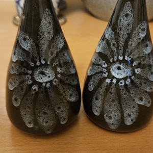 Könnte beinhalten: Zwei tropfenförmige Keramikstreuer mit glänzend dunkelbrauner Oberfläche. Jedes Stück zeigt ein weißes, punktiertes Blumenmuster, das von der Mitte ausstrahlt. Die Streuer haben eine abgerundete Basis und laufen oben spitz zu.