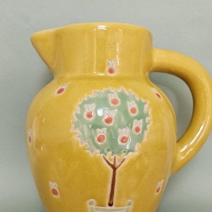 May include: Yellow ceramic jug with a handle and spout. The jug features a hand-painted design of a tree with red fruit and a green crown. The base has a striped pattern in red and white.