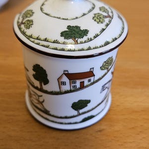 Op de afbeelding: Witte keramische pot met deksel, versierd met een landschap met een huis en bomen. De pot heeft een afgeronde basis en een conische knop op het deksel. Het ontwerp is in groen, bruin en wit.