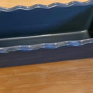 May include: A navy blue rectangular ceramic baking dish with a wavy, scalloped edge. The dish is empty and sits on a light brown wooden surface. This kitchenware is ideal for baking bread, cakes, or casseroles.