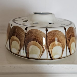 May include: A round ceramic flower frog with a white base and a repeating brown and tan arched pattern around the exterior. The top surface features a central opening surrounded by a series of smaller circular holes for arranging flower stems.