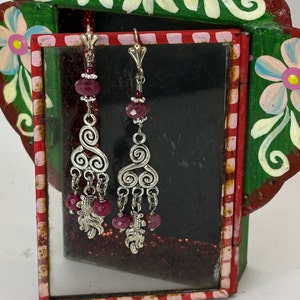 Puede incluir: Un par de pendientes largos y colgantes con filigrana plateada y cuentas de piedras preciosas rojas vibrantes. Los pendientes se exhiben frente a un marco decorativo pintado a mano con detalles florales en rosa, azul y blanco.