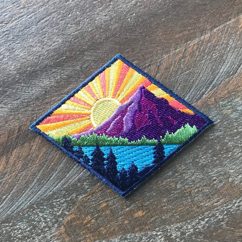 K&ouml;nnte beinhalten: Diamantf&ouml;rmiger gestickter Patch mit einer farbenfrohen Bergszene. Die Szene zeigt einen violetten Berg mit einer aufgehenden gelben Sonne dahinter, einen blauen See vor dem Berg und eine Reihe von dunkelgr&uuml;nen B&auml;umen im Vordergrund.