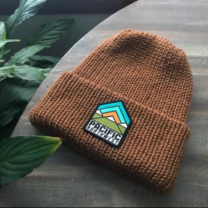 K&ouml;nnte beinhalten: Eine braune Strickm&uuml;tze mit einem schwarzen Aufn&auml;her, auf dem "Pacific Northwest" steht und ein Gebirge mit gr&uuml;nen, orangen und blauen Akzenten abgebildet ist.