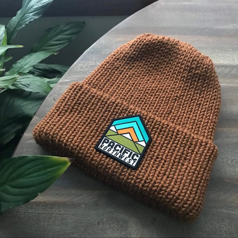 K&ouml;nnte beinhalten: Eine braune Strickm&uuml;tze mit einem schwarzen Aufn&auml;her, auf dem "Pacific Northwest" steht und ein Gebirge mit gr&uuml;nen, orangen und blauen Akzenten abgebildet ist.