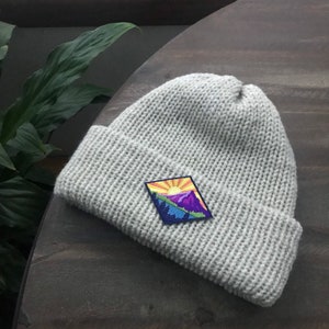 K&ouml;nnte beinhalten: Eine graue Strickm&uuml;tze mit einem bunten Patch, der eine Bergszene zeigt. Der Patch zeigt eine gelbe Sonne, lila Berge und gr&uuml;ne B&auml;ume.