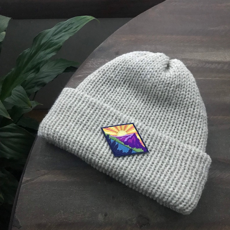 K&ouml;nnte beinhalten: Eine graue Strickm&uuml;tze mit einem bunten Patch, der eine Bergszene zeigt. Der Patch zeigt eine gelbe Sonne, lila Berge und gr&uuml;ne B&auml;ume.