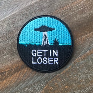 May include: A round black and blue embroidered patch with a UFO and the text "GET IN LOSER" in white.