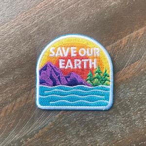 K&ouml;nnte beinhalten: Ein farbenfroher gestickter Aufn&auml;her mit einer Sonnenuntergangsszene und dem Text "SAVE OUR EARTH". Der Aufn&auml;her zeigt eine violette Bergkette, gr&uuml;ne B&auml;ume und blaues Wasser.