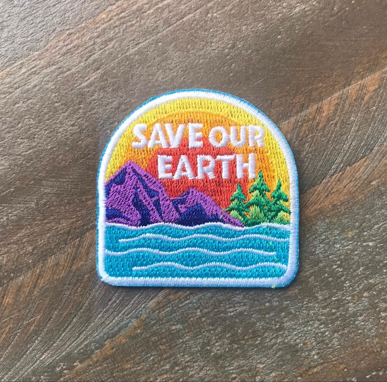 K&ouml;nnte beinhalten: Ein farbenfroher gestickter Aufn&auml;her mit einer Sonnenuntergangsszene und dem Text "SAVE OUR EARTH". Der Aufn&auml;her zeigt eine violette Bergkette, gr&uuml;ne B&auml;ume und blaues Wasser.