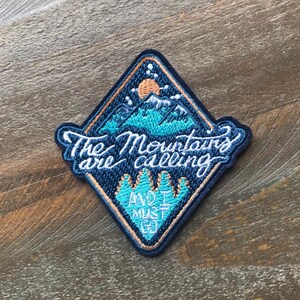 K&ouml;nnte beinhalten: Ein blauer und orangefarbener gestickter Aufn&auml;her mit einer Bergszene und dem Text "The Mountains are calling and I must go."