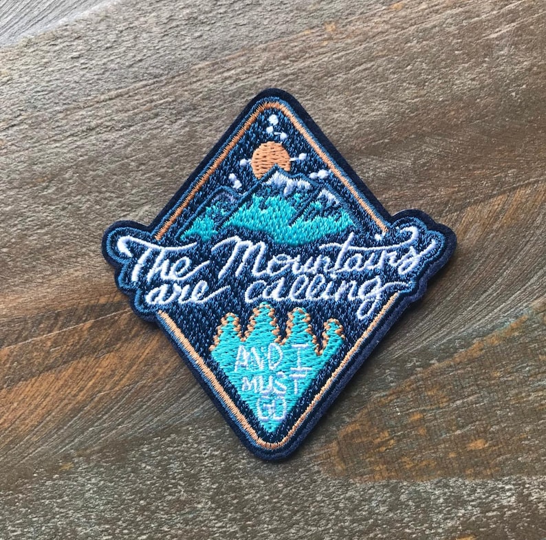 K&ouml;nnte beinhalten: Ein blauer und orangefarbener gestickter Aufn&auml;her mit einer Bergszene und dem Text "The Mountains are calling and I must go."