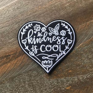 K&ouml;nnte beinhalten: Schwarzer herzf&ouml;rmiger Patch mit wei&szlig;er Stickerei, auf dem "kindness is cool" steht, umgeben von wei&szlig;en Blumenmustern.