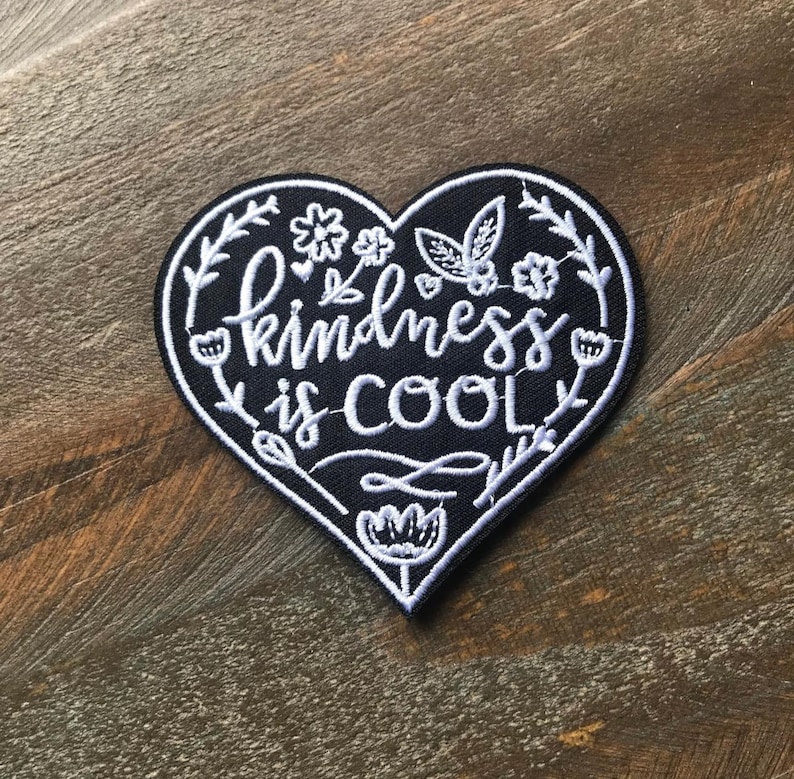 K&ouml;nnte beinhalten: Schwarzer herzf&ouml;rmiger Patch mit wei&szlig;er Stickerei, auf dem "kindness is cool" steht, umgeben von wei&szlig;en Blumenmustern.