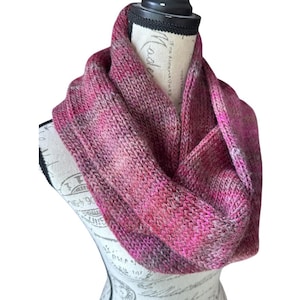 May include: A pink and purple infinity scarf made of knitted yarn. The scarf is displayed on a mannequin head. The color palette includes shades of pink, purple, and gray, creating a warm and cozy accessory.