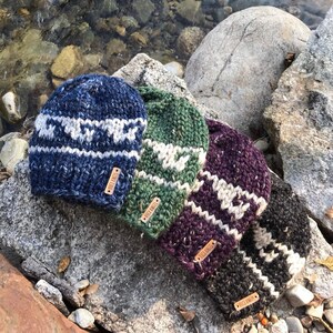 Op de afbeelding: Vier gebreide wollen mutsen met een wit en grijs patroon. De mutsen liggen in een rij op een rotsachtig oppervlak. Elke muts heeft een leren label met de tekst "Folk Knits" erop.