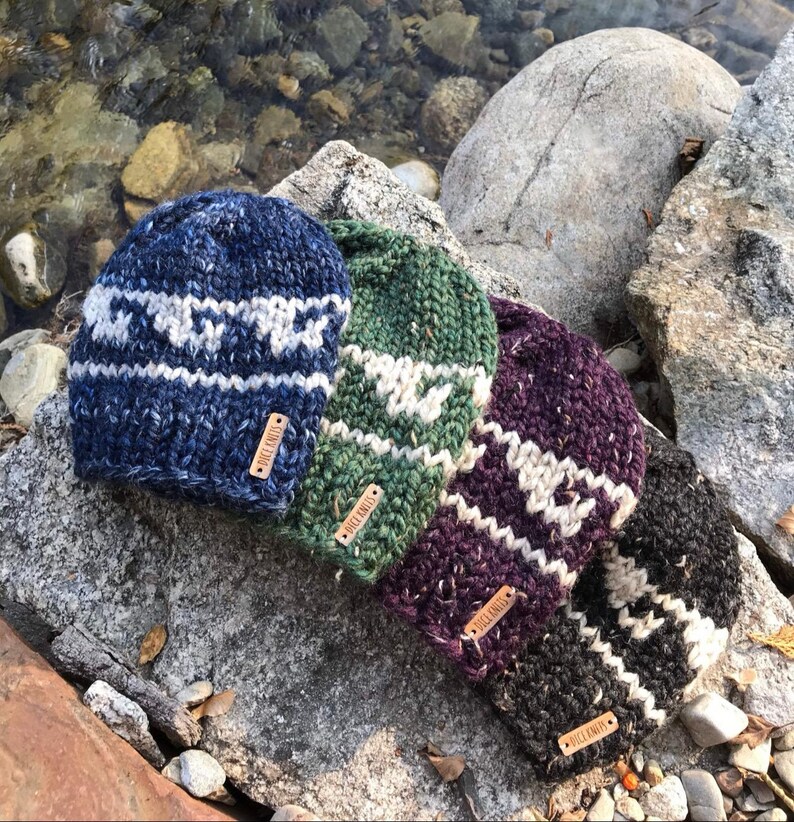 Op de afbeelding: Vier gebreide wollen mutsen met een wit en grijs patroon. De mutsen liggen in een rij op een rotsachtig oppervlak. Elke muts heeft een leren label met de tekst "Folk Knits" erop.