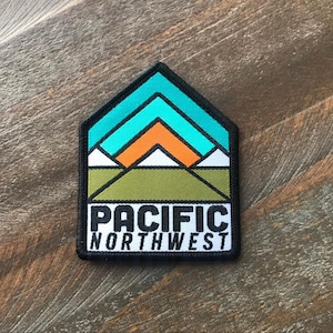 K&ouml;nnte beinhalten: Ein schwarzer und wei&szlig;er Patch mit einer Grafik einer Bergkette mit einem t&uuml;rkisfarbenen Himmel und einer orangefarbenen Sonne. Der Text "PACIFIC NORTHWEST" ist in Schwarz unter der Grafik gedruckt.